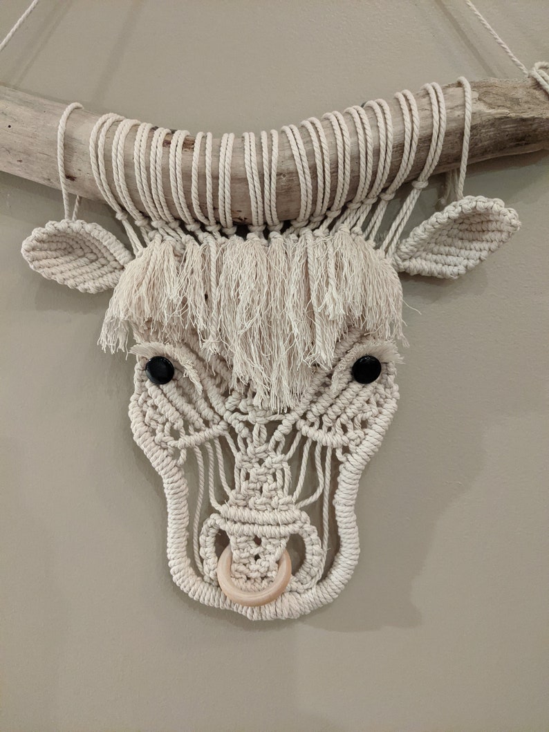 May include: A white macrame bull's head wall hanging with black button eyes and a wooden ring nose.