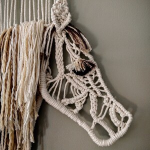 May include: A white macrame wall hanging in the shape of a horse's head. The horse's mane is made of brown and white yarn.