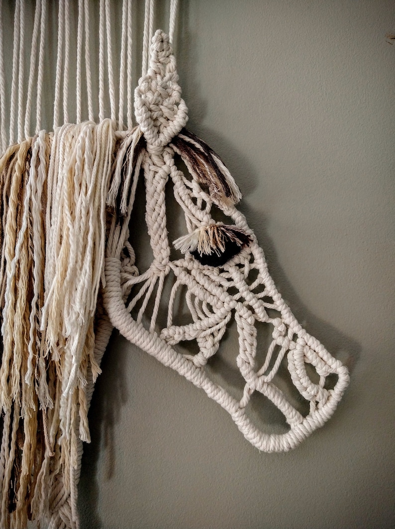 May include: A white macrame wall hanging in the shape of a horse's head. The horse's mane is made of brown and white yarn.