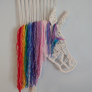 May include: A white macrame unicorn wall hanging with a rainbow coloured yarn mane. The unicorn is made with white cotton cord and has a detailed face and horn. The rainbow mane is made with a variety of colours including red, orange, yellow, green, blue, indigo, and violet.