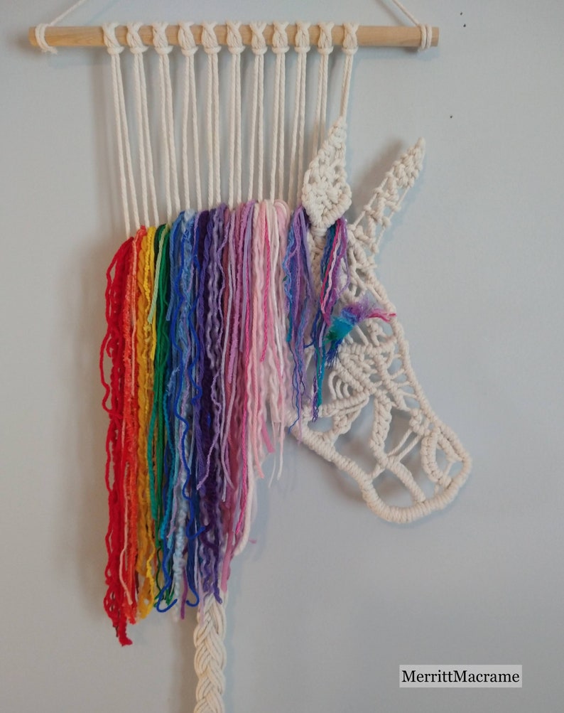 May include: A white macrame unicorn wall hanging with a rainbow coloured yarn mane. The unicorn is made with white cotton cord and has a detailed face and horn. The rainbow mane is made with a variety of colours including red, orange, yellow, green, blue, indigo, and violet.