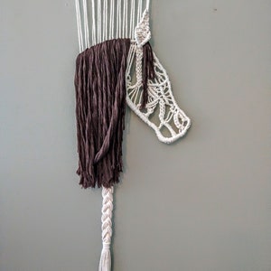 May include: A white macrame wall hanging with a brown fringe and a braided tail. The macrame is in the shape of a horse's head.