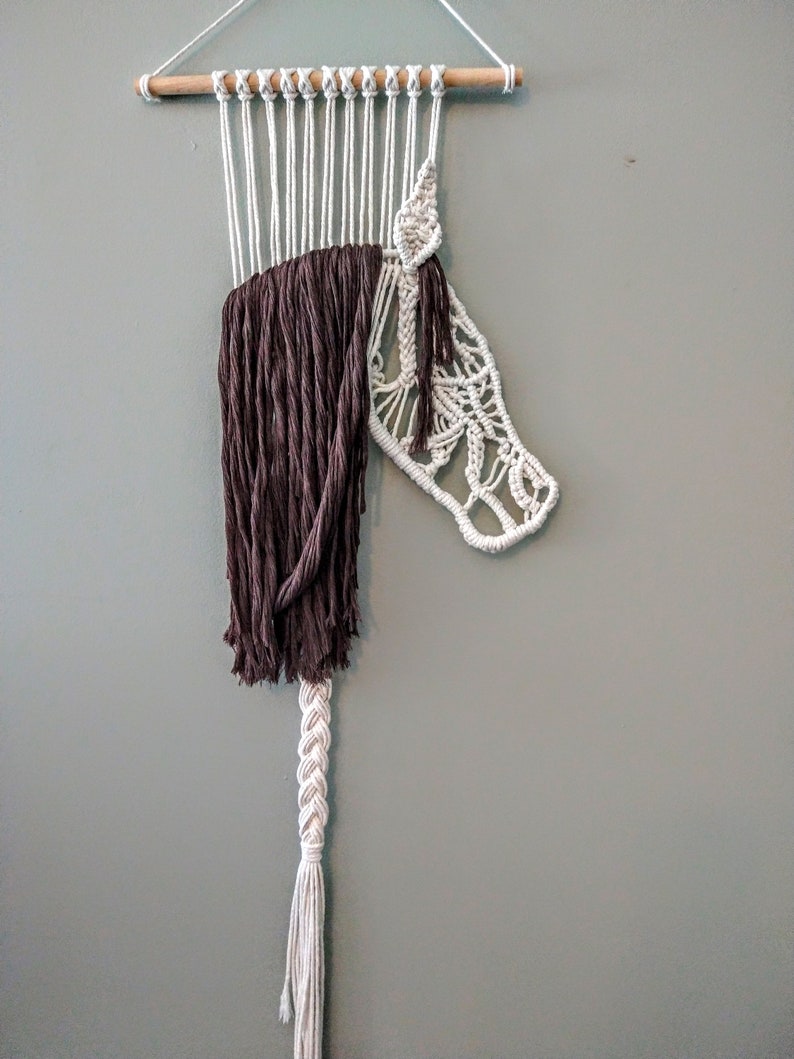 May include: A white macrame wall hanging with a brown fringe and a braided tail. The macrame is in the shape of a horse's head.