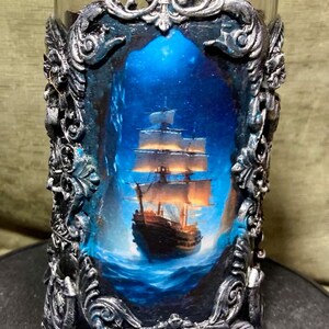 Op de afbeelding: Een heldere glazen kandelaar met een decoratief zilveren frame. Het frame omringt een schilderij van een schip dat op een blauwe zee vaart onder een sterrenhemel. De zeilen van het schip worden verlicht door warm licht.