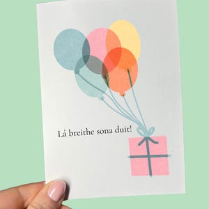 May include: A greeting card with a pastel colour scheme. The card features a cluster of balloons in blue, pink, orange, and yellow, tied to a pink gift box. The text "Lá breithe sona duit!" is printed on the card.