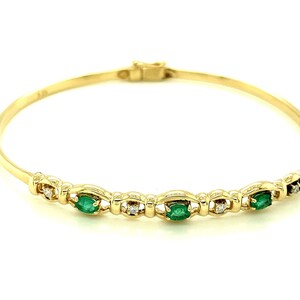 Könnte beinhalten: Ein goldener Armreif mit Smaragd- und Diamantakzenten. Das Armband hat eine Reihe von ovalen Smaragden, die zwischen kleinen Diamanten gesetzt sind. Der Verschluss ist oben sichtbar. Das Armband ist vor weißem Hintergrund.