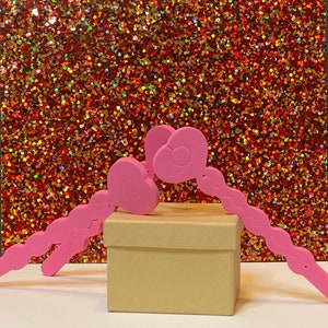 May include: A light brown cardboard box is topped with a pink decorative item. The pink item has a heart-shaped design with a wavy stem. The background is a glittery, multi-colored surface.
