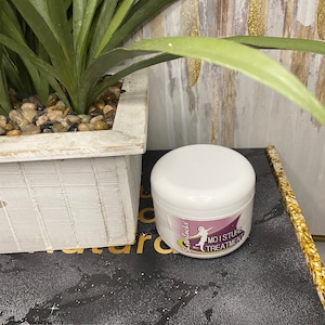 May include: A white jar of moisture treatment with a label featuring a woman silhouette and the text "Moisture Treatment". The jar is on a black surface next to a white wooden planter with a green plant.