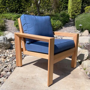 May include: A wooden outdoor chair with a blue cushion. The chair has a simple, modern design with a rectangular frame and a wide seat. The chair is sitting on a concrete patio.