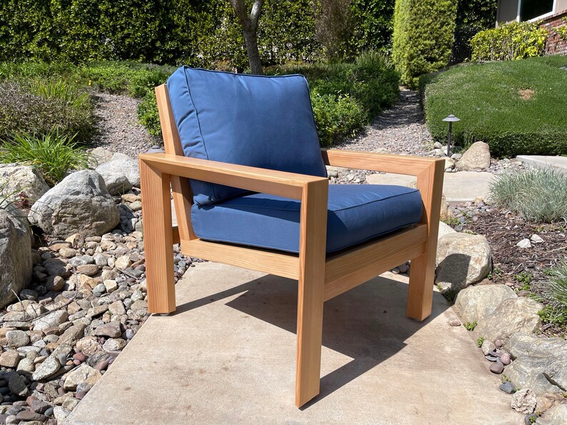 May include: A wooden outdoor chair with a blue cushion. The chair has a simple, modern design with a rectangular frame and a wide seat. The chair is sitting on a concrete patio.
