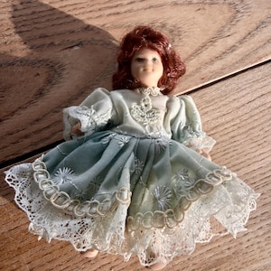 May include: A miniature doll with auburn hair and a pale complexion. The doll is dressed in a light blue dress with white lace trim and a detailed bodice. The doll is posed on a wooden surface.