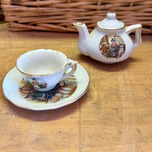 May include: A miniature porcelain tea set, comprising a small teapot and a teacup with a saucer. The set is white with gold edging and a colourful illustration of a person. The teapot has a lid and a handle. The teacup sits on a matching saucer.