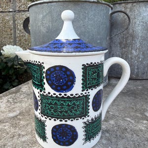 May include: A white ceramic jug with a lid, featuring a handle and spout. The jug is decorated with a repeating pattern of green and blue geometric shapes. The lid is white with a blue patterned design.