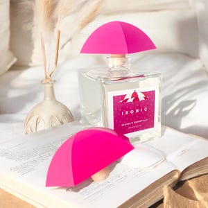 Op de afbeelding: Een heldere glazen fles met een roze paraplu-vormige stop. De fles is gelabeld met "IRONIC RACHEL'S ESSENTIALS 200ml/6.7 FL OZ".