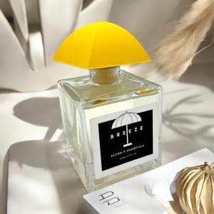 Może przedstawiać: Przezroczysta szklana butelka z żółtą pokrywką w kształcie parasola. Butelka jest oznaczona etykietą "BREEZE RACHEL'S ESSENTIALS 200ml/6.76 FL OZ".