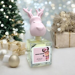 Pode incluir: Um tampão de garrafa de rena rosa com um design de rena festivo no rótulo. O rótulo diz "Holiday Edition" e "Diffuser Oil".