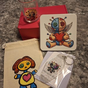 May include: A clear shot glass with a voodoo doll graphic, a square coaster featuring a winged voodoo doll, a beige drawstring pouch with a voodoo doll illustration, and a glittery motel-style keychain with the text MAGENTA BEJE and a voodoo doll.