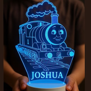 Könnte beinhalten: Blaues LED-Nachtlicht mit einem Cartoon-Zug-Design und dem Namen "JOSHUA" beleuchtet. Der Zug befindet sich auf Gleisen und stößt Rauch aus. Das Licht steht auf einem weißen Sockel.
