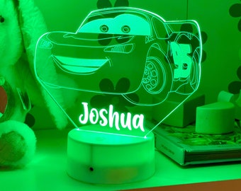 May include: A green light-up nightlight with a cartoon image of a red race car and the name "Joshua" in white. The nightlight is on a white base and is sitting on a shelf.