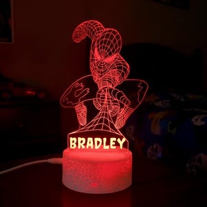 May include: A red LED night light featuring a flat, laser-etched acrylic silhouette of a superhero in a crouching pose. The base is cylindrical with a cracked texture, and the name BRADLEY is illuminated in red letters at the bottom of the figure.