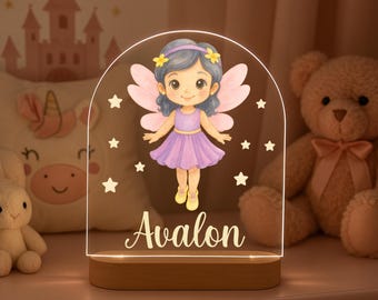 May include: A decorative night light featuring a cartoon fairy in a purple dress and wings, with the name "Avalon" illuminated in white. The light sits on a wooden base and is surrounded by star-shaped accents.
