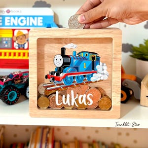 May include: A wooden money box with a clear front featuring a cartoon image of Thomas the Tank Engine. The name "Lukas" is written in white script below the train. A hand is dropping a coin into the slot.