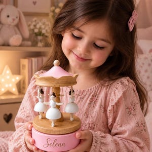 May include: A wooden musical carousel featuring three white ballerina figurines with dark hair, a natural wood base, and a pink roof. The cylindrical pink base is personalized with the name Selena in a cursive script font, centered on the front.