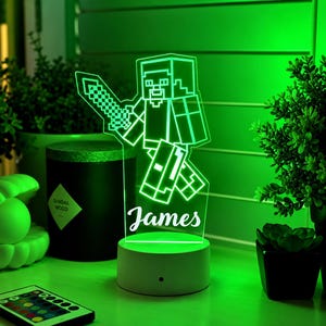 May include: A custom LED night light featuring a blocky, pixelated character holding a sword, illuminated in bright green. The clear acrylic panel displays the name James in a white script font, mounted on a circular white base with a power button.