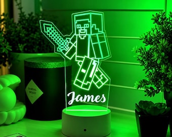 May include: A custom LED night light featuring a blocky, pixelated character holding a sword, illuminated in bright green. The clear acrylic panel displays the name James in a white script font, mounted on a circular white base with a power button.