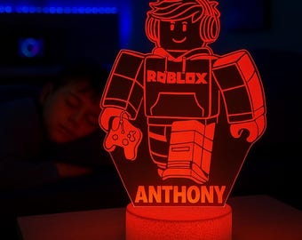 May include: A red LED night light featuring a Roblox character graphic wearing a hoodie and headphones while holding a game controller. The design includes the text Roblox on the chest and the name Anthony at the bottom, set on a circular base.