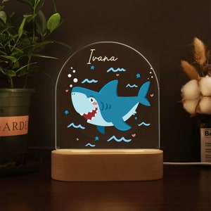 May include: A blue shark with a toothy grin is featured on a clear acrylic night light with a wooden base. The name "Ivana" is written in gold lettering at the top of the night light.