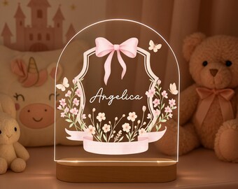 Pode incluir: Uma luz noturna personalizada com o nome "Angelica" em escrita cursiva. O painel de acrílico transparente apresenta um laço rosa, detalhes florais e uma moldura decorativa. A base é de madeira. A luz está acesa.