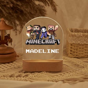 以下が含まれることがあります： Minecraftのキャラクターデザインと「MADELINE」という名前がピクセルフォントで印刷された透明なアクリル製のナイトライト。ライトは木製ベースです。