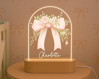 Pode incluir: Uma luz noturna decorativa com base de madeira e painel acrílico transparente. O painel apresenta um laço rosa e um design floral, com o nome "Charlotte" em escrita cursiva. A luz emite um brilho quente, ideal para um quarto.