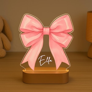 Op de afbeelding: Een roze strikvormig nachtlampje met de naam "Ella" in cursief schrift. De strik is verlicht en staat op een houten voet. Het licht is ontworpen als decoratief item.
