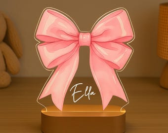 Pode incluir: Uma luz noturna em forma de laço rosa com o nome "Ella" em escrita cursiva. O laço é iluminado e fica sobre uma base de madeira. A luz foi projetada para ser um item decorativo.