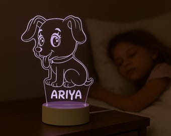 May include: An illuminated LED night light featuring a cartoon dog design, with the name "ARIYA" displayed. The light sits on a bedside table, next to a sleeping child. The dog is white with a black outline.