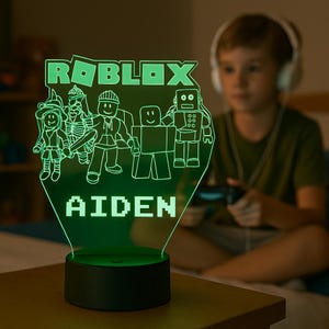 May include: A glowing green LED night light featuring a flat acrylic panel with the Roblox logo, various character figures, and the name AIDEN in block letters. The light is mounted on a circular black base with a smooth, matte finish.