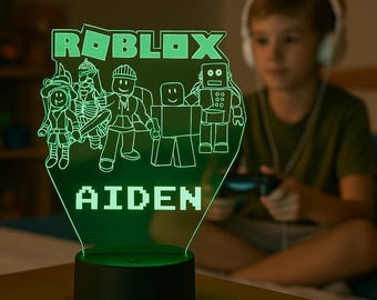 May include: A glowing green LED acrylic lamp featuring the Roblox logo and various character figures, with the name AIDEN printed in block letters at the bottom. The light is mounted on a circular black base and emits a bright neon green light.