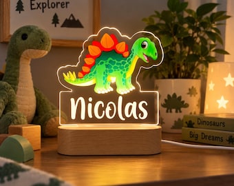 Könnte beinhalten: Ein personalisiertes LED-Nachtlicht mit einem bunten grünen Dinosaurier mit orangefarbenen Platten auf dem Rücken, montiert auf einem hellen Holzsockel. Die Acrylplatte zeigt den Namen Nicolas in weißer Schrift, beleuchtet durch ein warmes Licht.