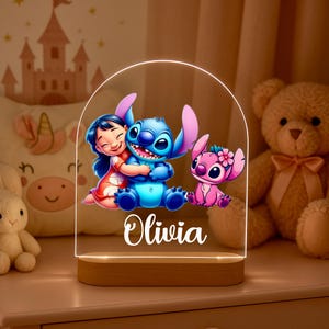 Peut inclure: Une veilleuse décorative avec les personnages Lilo & Stitch. La lampe a une base en bois et un panneau acrylique transparent avec les personnages et le nom "Olivia" illuminés. Les personnages sont dans des tons de bleu, rose et rouge.
