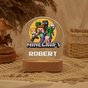 以下が含まれることがあります： Minecraftのキャラクターデザインと「ROBERT」という名前がピクセルフォントで印刷された白いアクリル製のナイトライト。ライトは木製のベースの上に置かれています。
