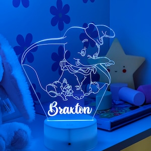 May include: A white night light with a blue glow features a line drawing of Dumbo the elephant with his mouse friend. The light is on a white base and the name "Braxton" is written below the image.