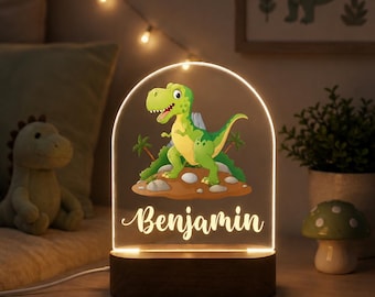 Puede incluir: Una luz nocturna de acrílico en forma de arco con un colorido Tyrannosaurus Rex de dibujos animados sobre una isla rocosa con palmeras. El nombre Benjamin está impreso en letra cursiva blanca debajo del dinosaurio. Base de madera oscura.