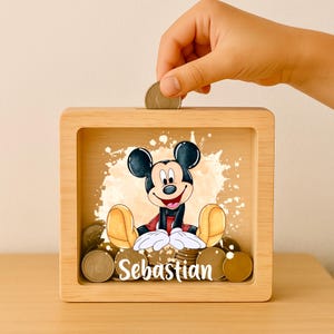 Può includere: Un salvadanaio in legno con un'illustrazione di Topolino sorridente. La scatola ha un fronte trasparente, una fessura per inserire le monete e il nome "Sebastian" stampato. La scatola è piena di monete e una mano sta inserendo un'altra moneta.