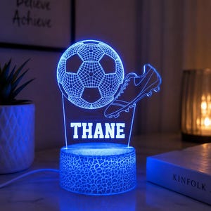 May include: A glowing blue LED night light featuring a flat acrylic panel with an etched soccer ball and a soccer cleat. The name THANE is printed in white capital letters below the ball. The light sits on a cylindrical base with a cracked texture.