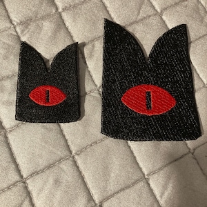 May include: Two black fabric patches with a red eye design. The patches are different sizes.