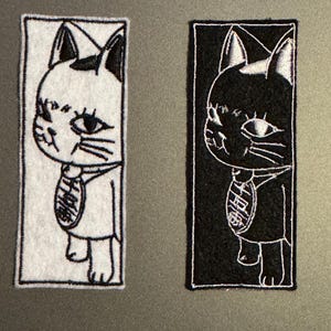 Könnte beinhalten: Zwei gestickte Bügelbilder mit einer Cartoon-Katze mit Halsband und Anhänger. Die Katze links ist weiß mit schwarzen Umrissen und die Katze rechts ist schwarz mit weißen Umrissen.