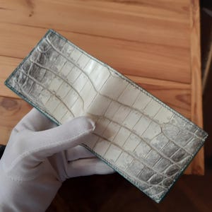 Puede incluir: Una cartera plegable con un patrón de piel de cocodrilo texturizado en color blanco roto y gris. La cartera tiene una forma rectangular con costuras visibles a lo largo de los bordes, mostrando una textura superficial escamosa distintiva.