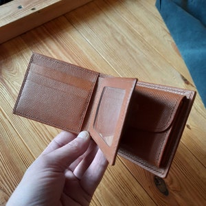 以下が含まれることがあります： IDカード用の透明なプラスチック窓が付いた茶色の革財布。財布は開いて半分に折り畳まれており、内側のポケットが見えています。